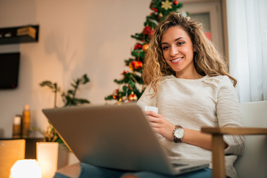 Eine junge Frau mit blondem lockigen Haar blick auf Ihren Laptop, der auf ihrem Schoß steht. In der linken Hand hält Sie eine Tasse, sie lächelt. Im Hintergrund ist ihr Zimmer zusehen, in dem auch ein Weihnachtsbaum steht.