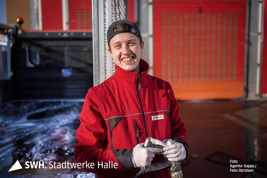 Ein junger Mann lächelt in die Kamera. Er steht vor einem Gebäude, blickt in die Kamera, trägt eine rote Jacke und weiße Handschuhe.