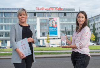 Das Foto wurde vor dem Stadtwerke-Gebäude aufgenommen und zeigt zwei Frauen, die in die Kamera lächeln. Die Frau auf der linken Seite hält eine Sonderpreis-Urkunde in der Hand, die Frau auf der rechten Seite eine gläserne Trophäe.